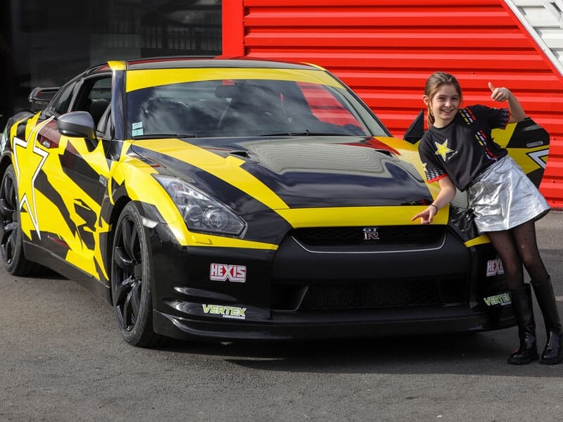 Une jeune fille pose souriante devant une Nissan GTR jaune et noire