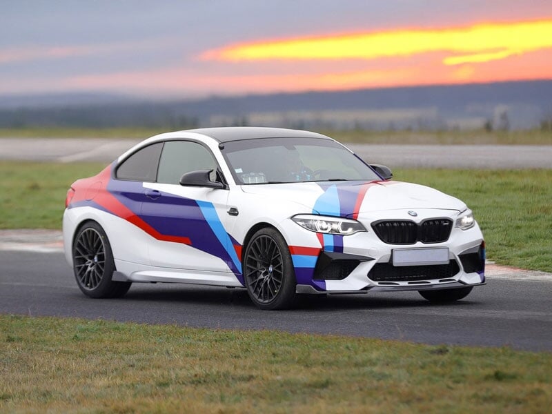 BMW M2 Compétition blanche avec un revêtement spécial aux motifs bleu, rouge et violet inspirés de la division Motorsport. Voiture sportive équipée de jantes en alliage noir, calandre double caractéri