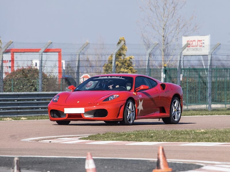 Ferrari F430 rouge sur circuit de Dijon Prenois lors d'un stage de pilotage pour enfant, sous un ciel clair.