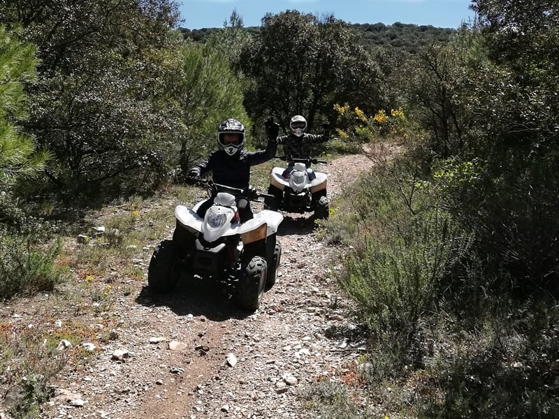 Deux personnes en quad sur un chemin de terre entouré de végétation dense sous un ciel clair.