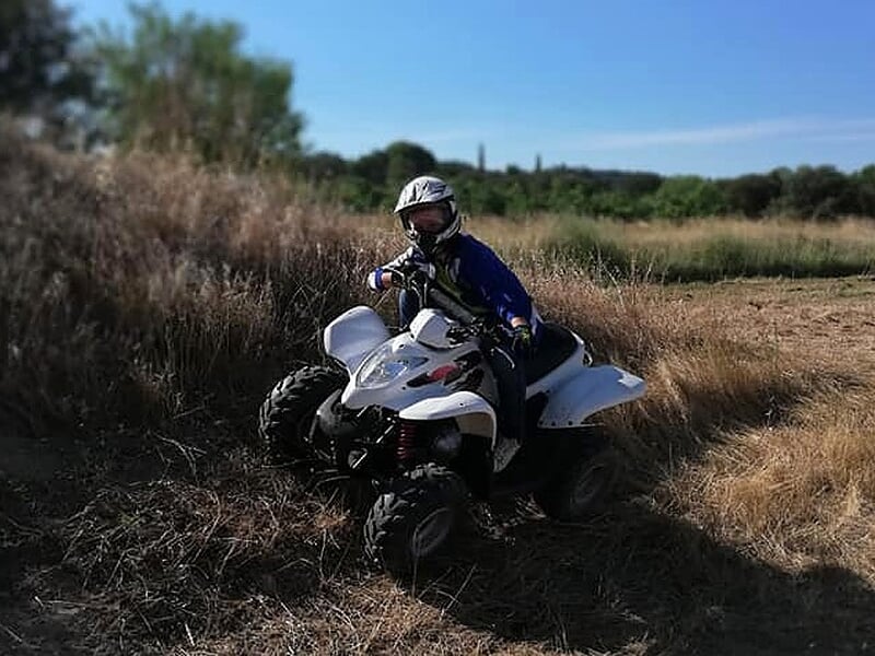 Enfant en tenue de protection sur un quad blanc, roulant sur un chemin entouré d'herbes sèches.