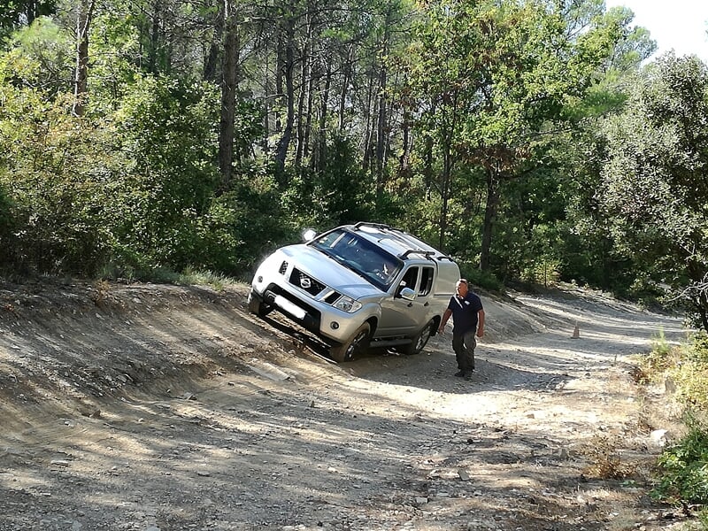 Véhicule 4x4 blanc en descente sur un chemin accidenté en forêt, avec une personne à côté.
