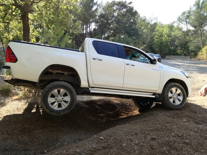 Camionnette blanche tout-terrain franchissant un obstacle en terrain boisé et accidenté.