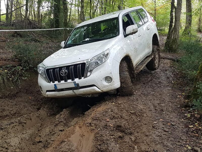 Véhicule 4x4 blanc boueux roulant sur un chemin forestier étroit et accidenté.