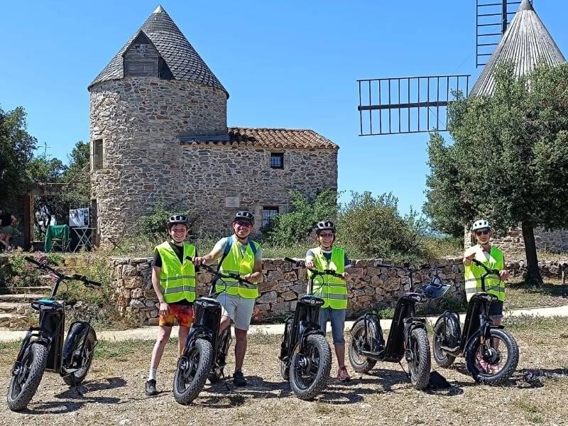 Groupe de cinq personnes en gilets jaunes posant devant un moulin à vent en pierre, tous avec des trottinettes électriques, sous un ciel bleu clair.