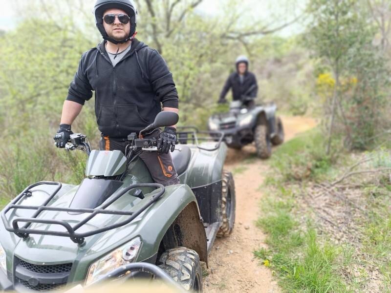 Homme en quad tout-terrain vert, vêtu de noir avec des lunettes de soleil et un casque, sur un chemin de terre bordé de végétation.