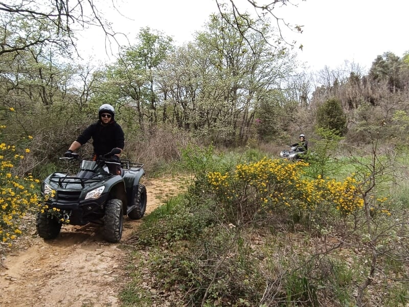 Deux hommes en quad tout-terrain, l'un au premier plan sur un chemin de terre, l'autre en arrière-plan, entourés de végétation dense et de fleurs jaunes.