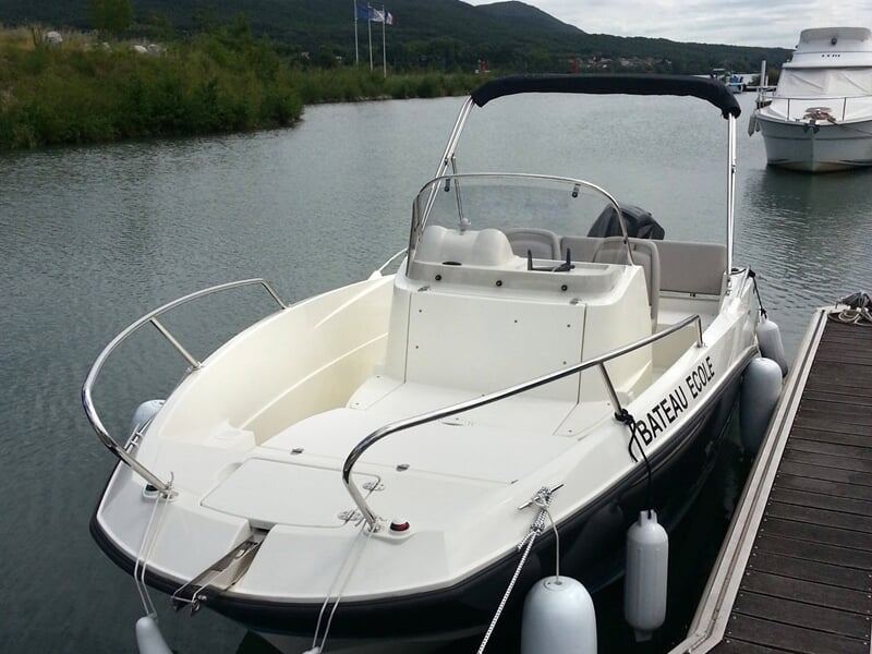 Petit bateau blanc amarré à un quai sur une rivière calme avec des collines en arrière-plan sous un ciel nuageux.