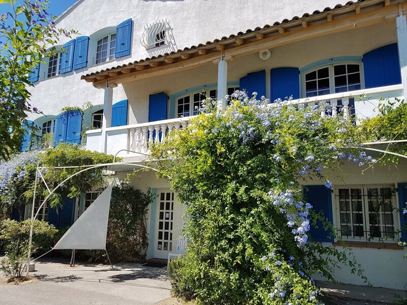 Maison blanche avec volets bleus, balcon et jardin fleuri sous un ciel clair et ensoleillé.