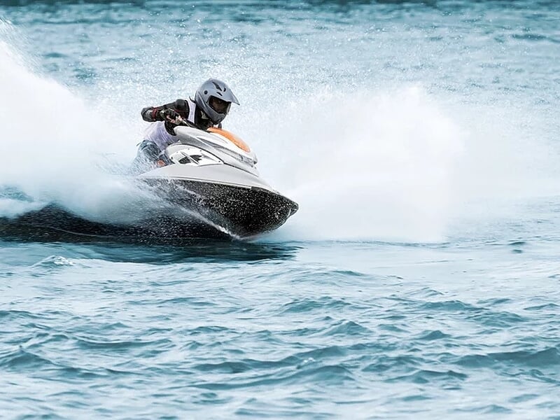 Conducteur en casque pilotant un jet-ski blanc et orange à vive allure, soulevant des gerbes d’eau sur une mer turquoise.