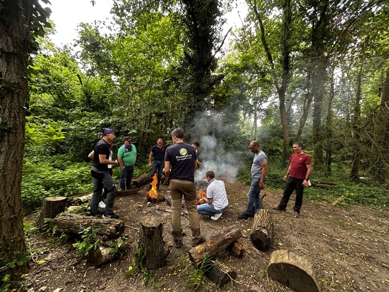 Groupe de personnes réunies autour d'un feu de camp dans une clairière boisée, ambiance conviviale en extérieur.