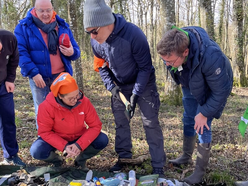 Groupe de personnes en tenue d'extérieur examinant du matériel posé sur une bâche dans un environnement boisé.