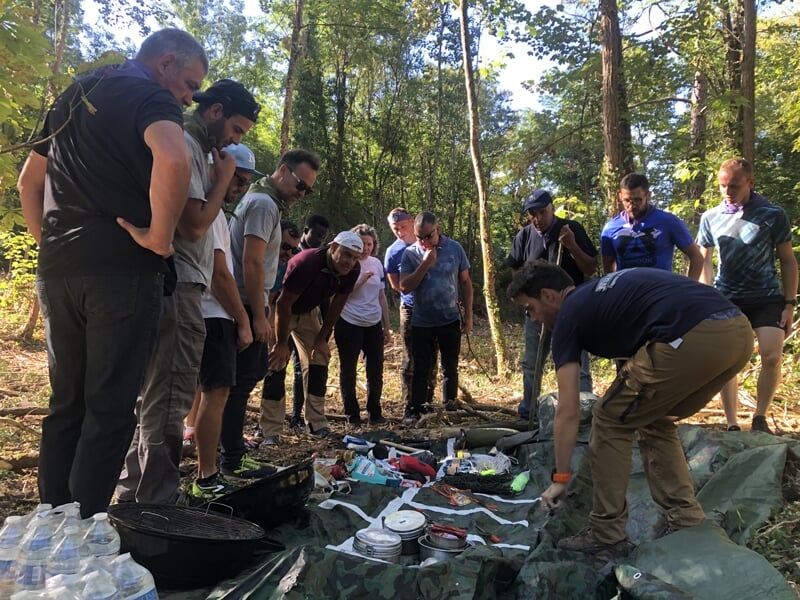 Groupe nombreux d'adultes attentifs autour d'un instructeur qui présente du matériel de survie en forêt.