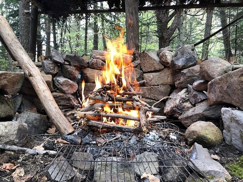 Feu de camp allumé dans un cercle de pierres avec un grillage au-dessus en forêt dense et boisée.