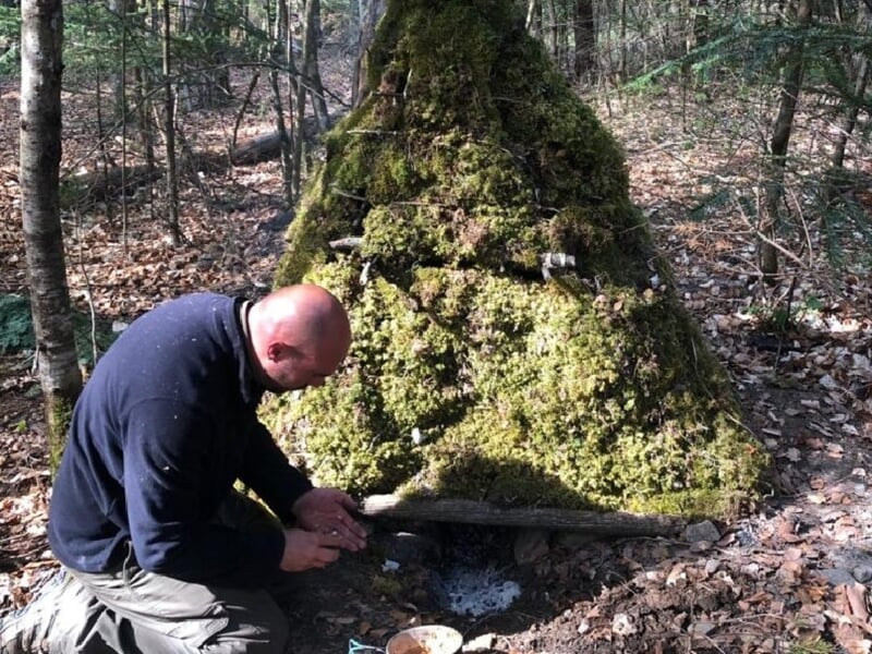 Homme agenouillé devant un abri en forme de tipi recouvert de mousse dans une forêt verdoyante.