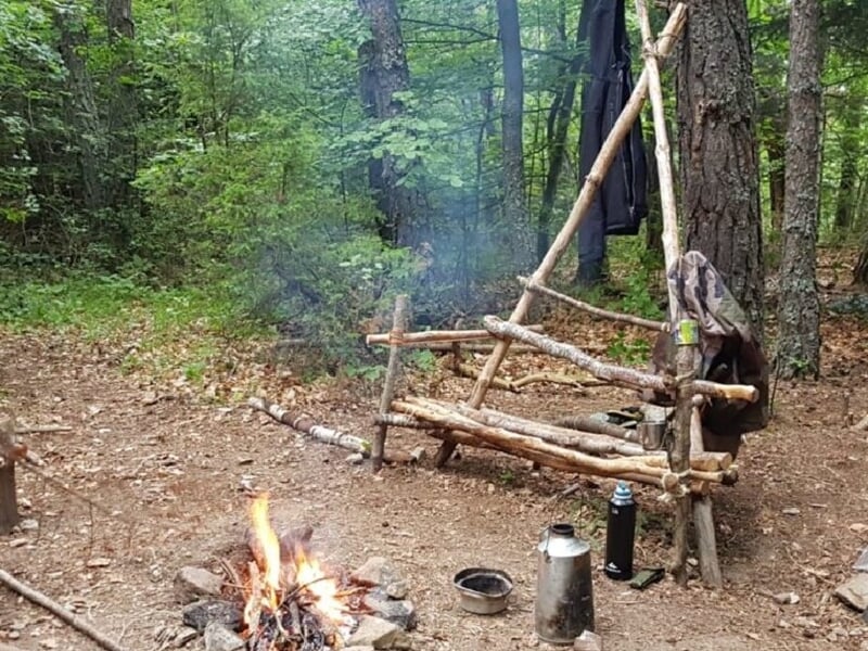 Campement en forêt avec un feu de camp, un banc en bois fait de branches et des objets posés au sol.