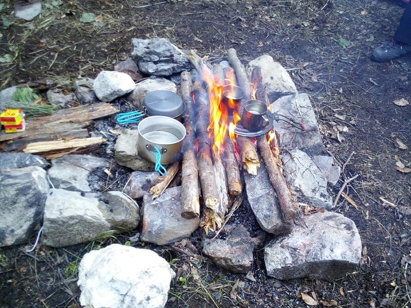 Feu de camp entouré de pierres avec des casseroles et du bois pour cuisiner en pleine nature.