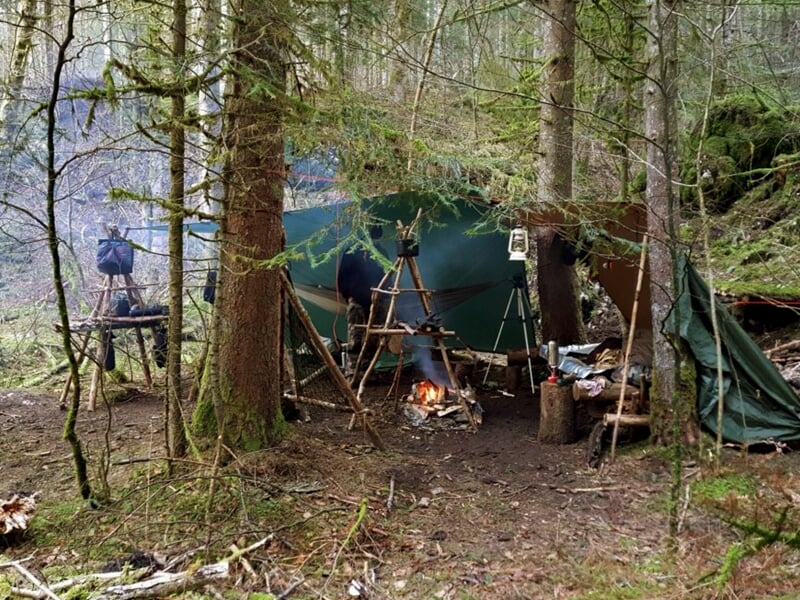 Campement de survie en forêt avec un feu de camp, une tente de fortune et du matériel suspendu.