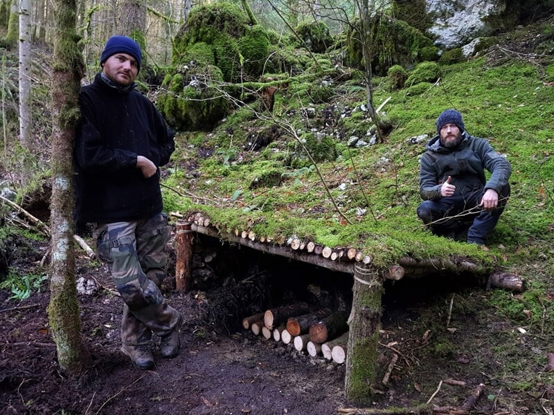 Deux hommes posant fièrement à côté d'un abri de survie construit avec des branches et recouvert de mousse.