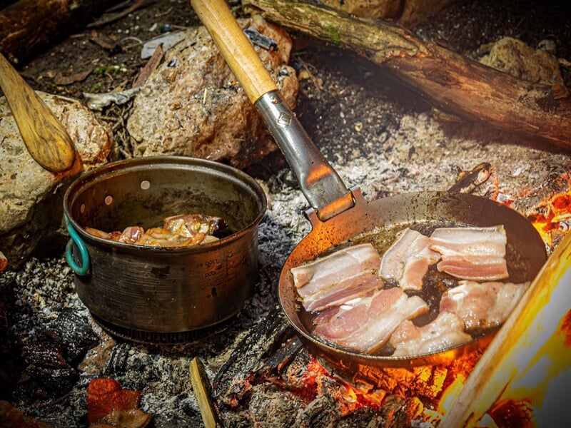 des tranches de lard et d'autres aliments cuisent sur un feu de camp en pleine nature