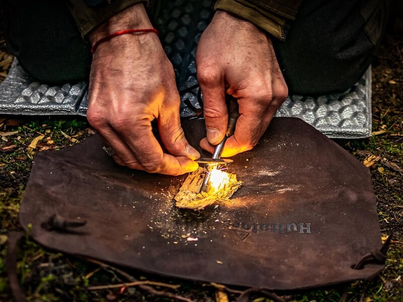 Mains d'une personne allumant un feu avec un briquet et un morceau de bois sec sur un tapis de sol.
