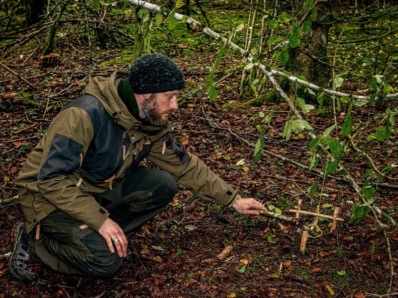 Homme accroupi dans la forêt, manipulant un piège en bois fabriqué à la main pour la survie.