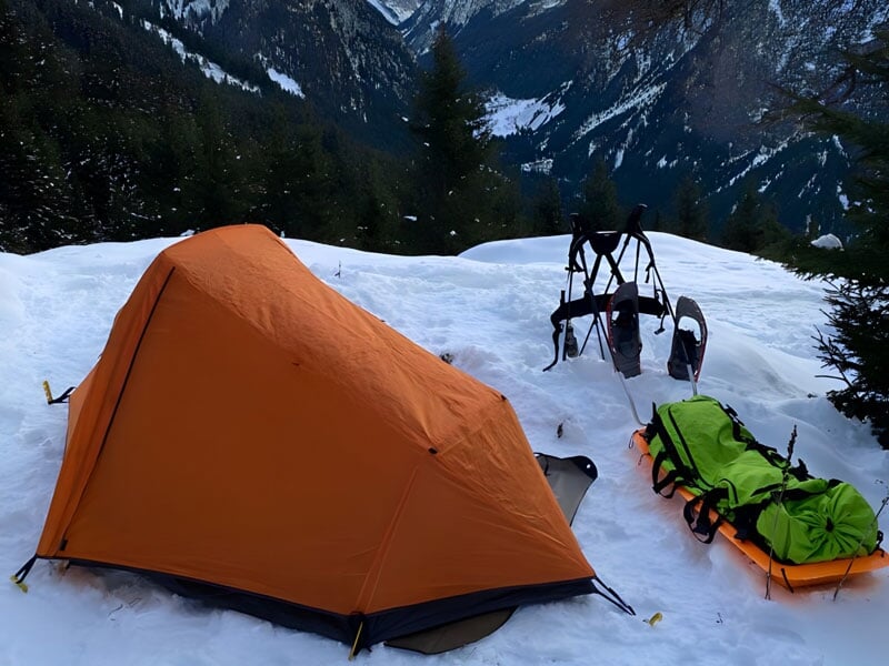 une tente de camping et une pulka sont installées dans le neige devant une vallée enneigée
