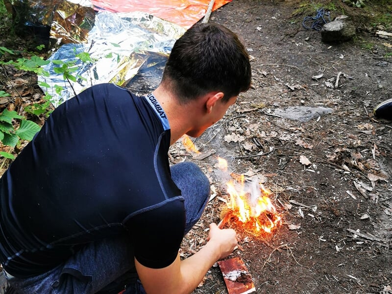 Un homme allumant un feu de camp dans une forêt avec une couverture de survie argentée en arrière-plan