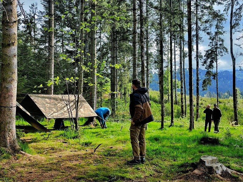 Randonneurs en forêt avec un abri monté, un dans le hamac et deux autres debout à discuter, avec des arbres et une clairière en arrière-plan lors d'un stage de survie bushcraft