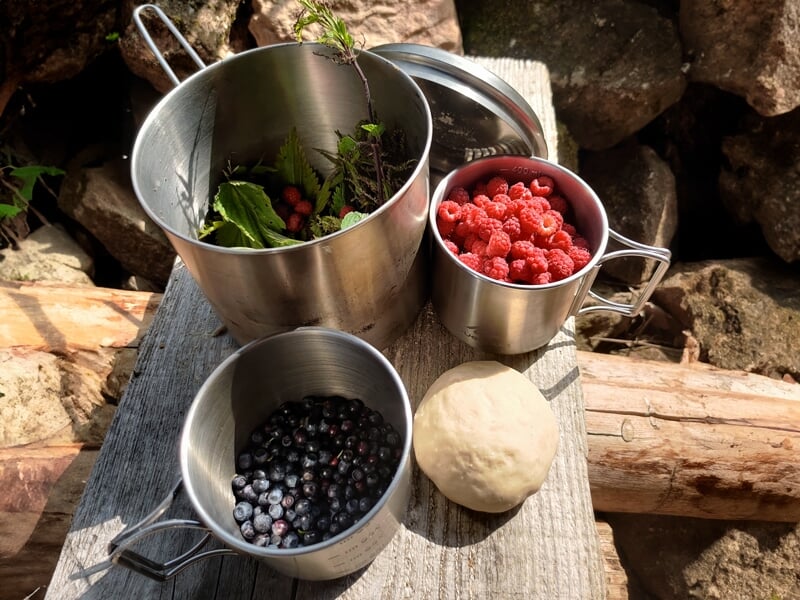 Trois récipients métalliques contenant respectivement des feuilles vertes, des framboises et des myrtilles, placés sur une planche de bois à côté d'une boule de pâte, prêts pour la cuisson
