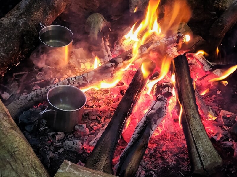 Feu de camp vibrant avec des flammes brillantes, des tisons rouges, et deux casseroles métalliques posées à côté, illustrant un moment de cuisson en pleine nature