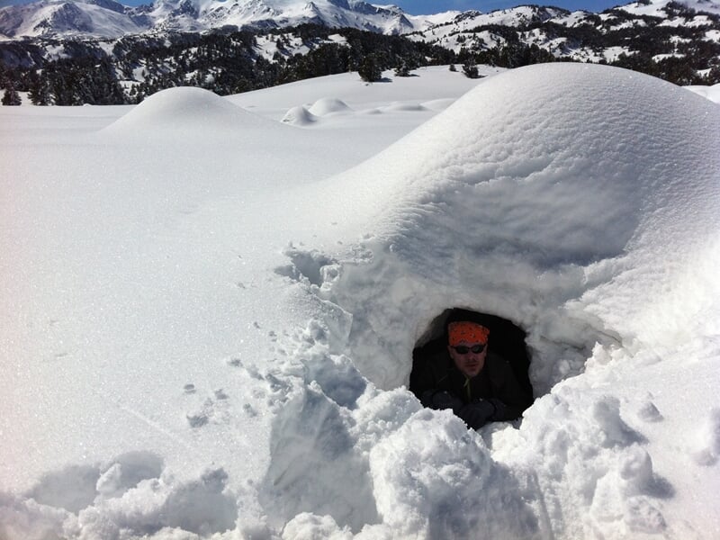 Personne sortant d'un abri creusé dans la neige avec un paysage montagneux enneigé en arrière-plan.
