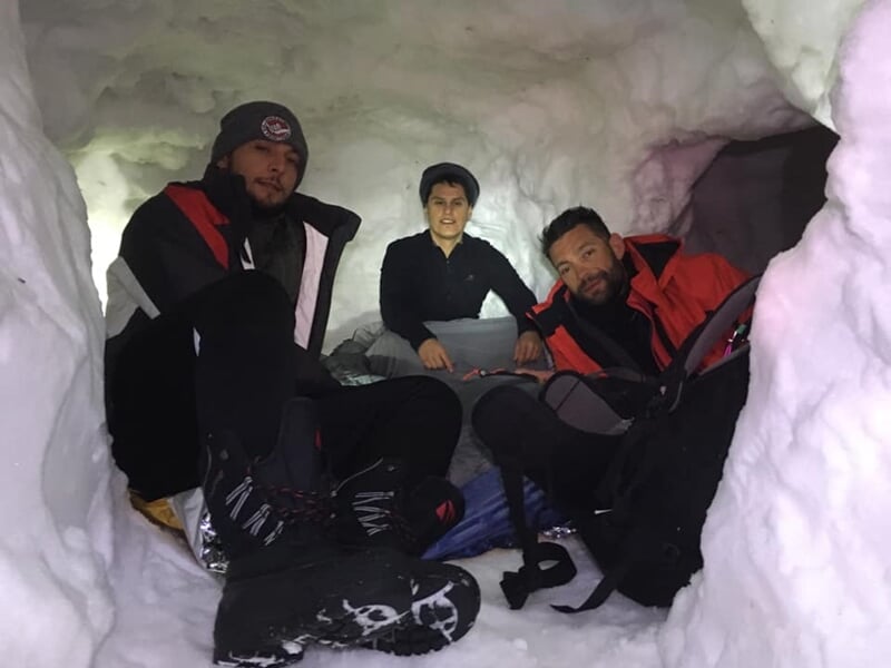 Trois personnes assises dans un abri de neige, souriantes et détendues, entourées de sacs et équipements.