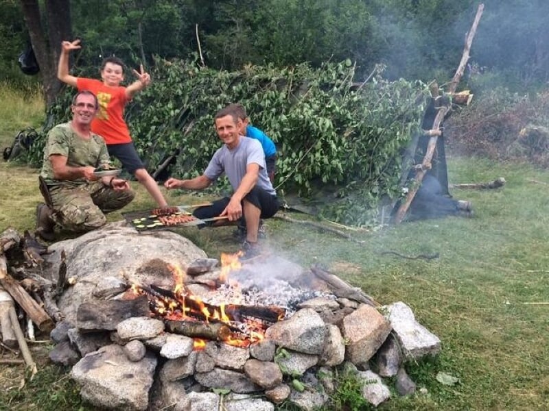 Trois personnes autour d'un feu de camp en plein air, cuisinant des aliments sur des bâtons dans un cadre naturel.