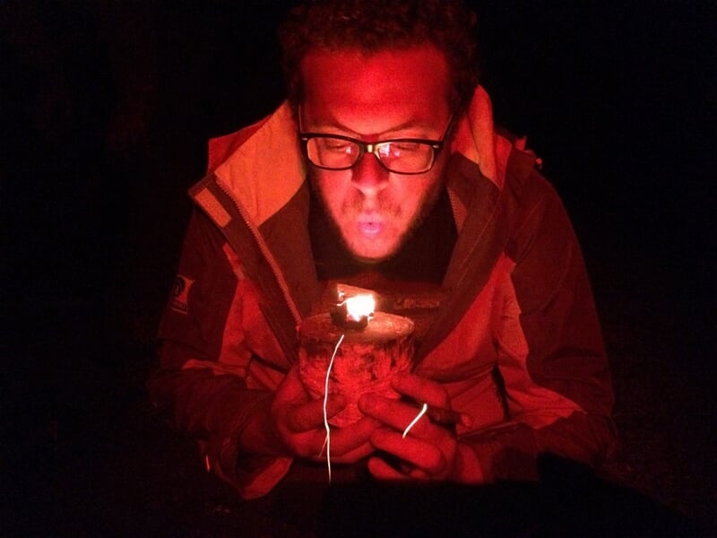 Homme soufflant sur un feu naissant dans l'obscurité, éclairé par la lumière rouge du feu.