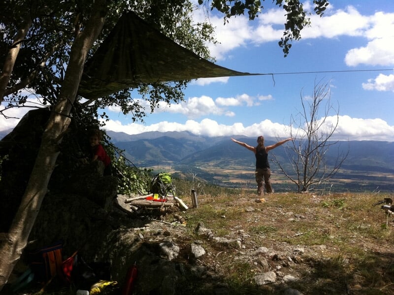 Personne debout sur un promontoire naturel, bras ouverts, avec une vue panoramique sur une vallée et montagnes.