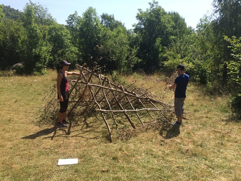 Deux personnes debout dans un champ, observant une structure en bois en forme de tipi partiellement construite.