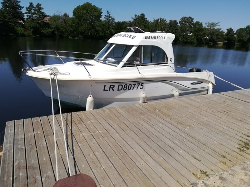Petit bateau école blanc amarré à un ponton en bois sur un plan d'eau calme avec des arbres en arrière-plan