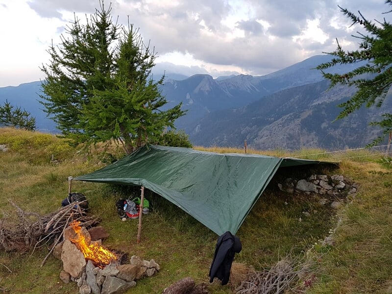 Abri de fortune en toile tendue dans un paysage montagneux avec un feu de camp allumé à proximité