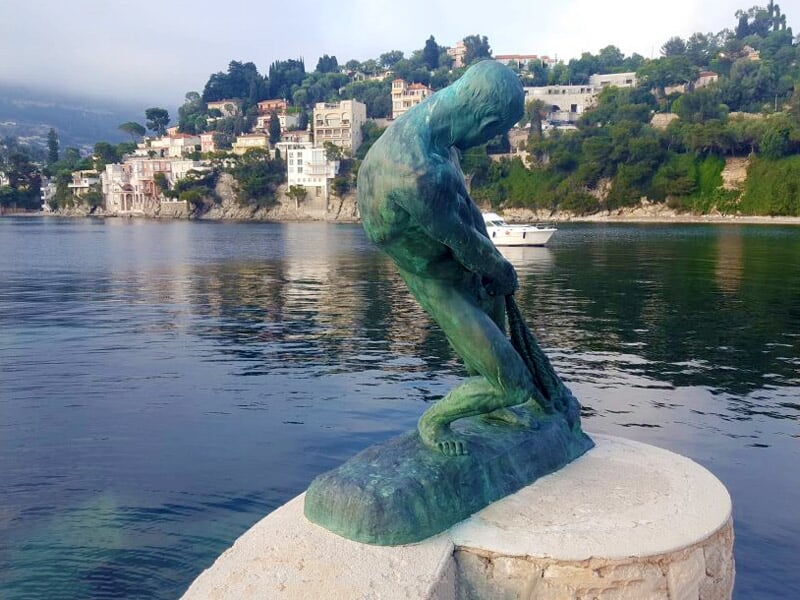Statue en bronze d'un homme penché sur un rocher au bord de l'eau avec des maisons en arrière-plan