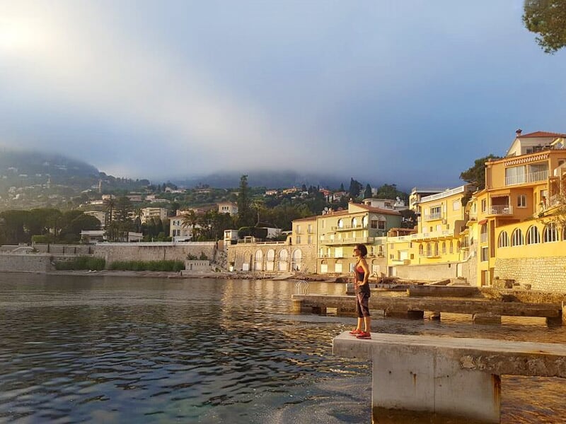 Personne debout sur un quai en bord de mer avec des maisons et collines en arrière-plan au coucher du soleil