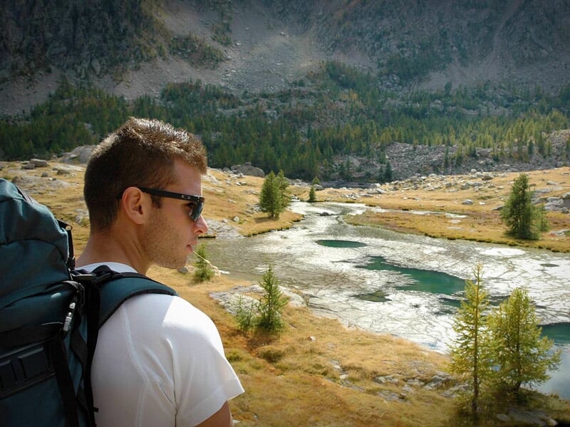 Homme avec sac à dos regardant un lac de montagne entouré de forêts et de sommets rocheux