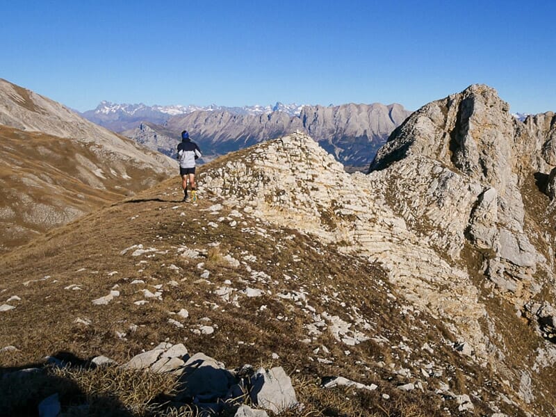 Un coureur en tenue sportive court sur un sentier de montagne rocheux avec un paysage alpin en arrière-plan.