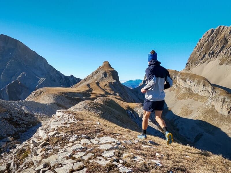 Un homme en tenue de trail court sur une crête étroite entourée de montagnes rocheuses et d'un ciel bleu.