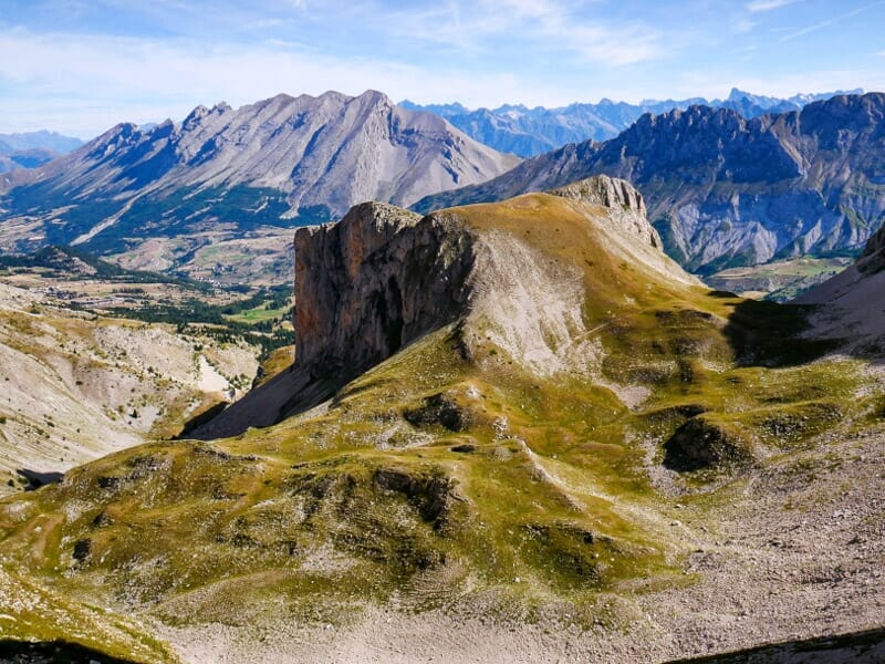 Vue panoramique sur une vallée de montagne avec des sommets rocheux et des prairies verdoyantes sous un ciel clair.