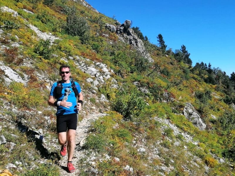 Coureur de trail en montagne sur un sentier entouré de végétation et de rochers sous un ciel bleu clair