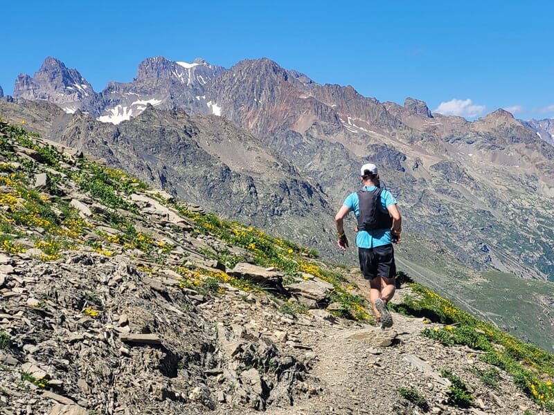 Un coureur en tenue sportive court sur un sentier de montagne rocailleux avec un paysage de sommets en arrière-plan.