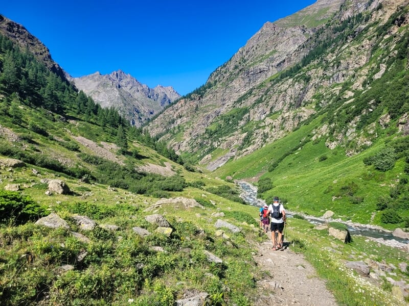 Deux randonneurs marchent sur un sentier entouré de végétation dense et de montagnes sous un ciel bleu.