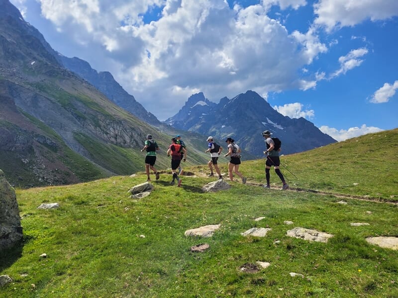Quatre coureurs en trail avancent sur une crête herbeuse avec des montagnes imposantes et un ciel nuageux.