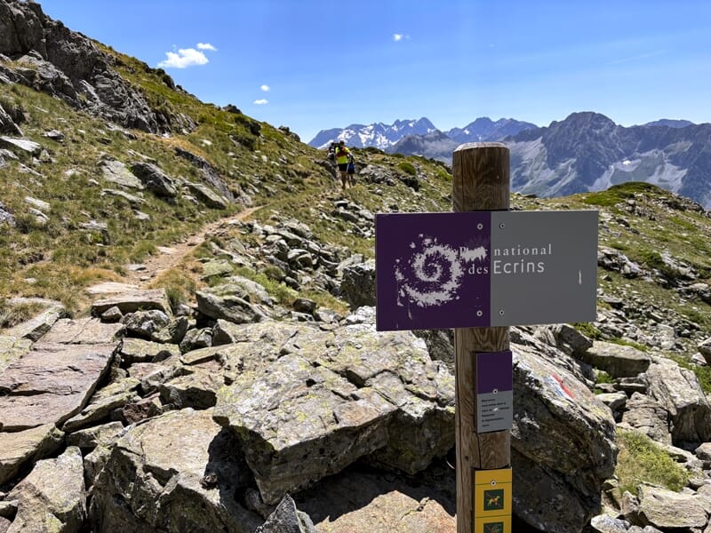 Panneau du parc national des Écrins sur un sentier de montagne rocheux avec un randonneur en arrière-plan.